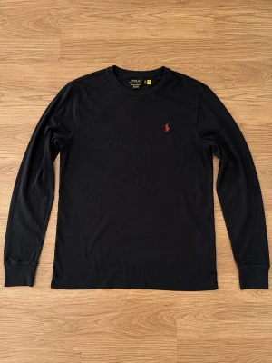 Svart långärmad topp från Polo Ralph Lauren - Svart långärmad topp från Polo Ralph Lauren med klassisk rund hals och rött broderat logomärke på bröstet. Tillverkad i mjuk bomull som känns skön mot huden. Perfekt för en clean och stilren look.