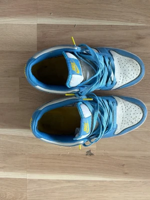 Nike Dunk Low blå och vita sneakers - Nike Dunk Low sneakers i blått och vitt med gula detaljer. Skorna har klassisk låg siluett, perforerad tåbox och blå swoosh på sidorna. Ovandelen är i läder och textil, med matchande blå skosnören och gul Nike-logga på plösen och hälen.