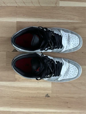 Air Jordan 1 Low Vit/Svart/Grå - Snygga Air Jordan 1 Low sneakers i vitt, svart och grått läder med svarta skosnören och röd logga på plösen. Klassisk svart swoosh på sidan och röd detalj på hälen. Perfekta för dig som gillar streetwear och vill sticka ut.