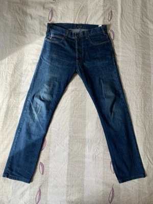 Blå raka jeans från Armani Jeans - Snygga blå jeans från Armani Jeans med klassisk femficksdesign och raka ben. Jeansen har subtila slitningar och kontrastsömmar, samt en metallknapp med logga. Perfekta för en stilren och avslappnad look.