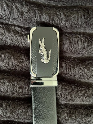 Svart bälte med silverdetaljer - Snyggt svart bälte från Lacoste med silverfärgat krokodilemblem på spännet. Bältet är gjort i skinn och har en stilren, modern design med präglat mönster på spännet. Perfekt accessoar för att lyfta din outfit.