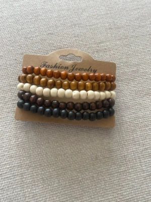 Set med pärlarmband i trä - Säljer ett set med fem elastiska armband från Fashion Jewelry, gjorda av runda träpärlor i olika färger: svart, mörkbrun, ljusbrun, beige och orangebrun. Perfekta att mixa och matcha för en cool och avslappnad stil.