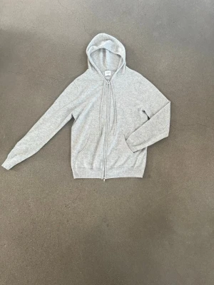Ljusgrå zip-hoodie med huva - En stilren ljusgrå cashmere hoodie från John heric med dragkedja och huva. Tröjan har långa ärmar, ribbade muddar och dragsko i huvan. Perfekt för en avslappnad look och enkel att matcha med andra plagg.