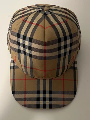 Beige keps med klassiskt rutmönster - Säljer en snygg keps från Burberry med deras ikoniska beige rutmönster i svart, vitt och rött. Kepsen har böjd skärm och justerbar baksida. Perfekt för dig som vill ha en trendig accessoar med lyxig känsla.