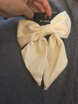 Krämvit stor hårrosett från Lyko - Säljer en stor och elegant hårrosett i krämvitt tyg från Lyko. Rosetten har en lyxig satinliknande finish och är perfekt för att ge frisyren en extra touch. Enkel att fästa i håret och passar till många olika stilar.