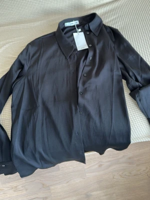 Svart silkesblandad skjorta från InWear - Säljer en svart skjorta från InWear med klassisk krage och knappar framtill. Skjortan är långärmad och har en elegant, slät finish i silkesblandat material som ger en lyxig känsla. Perfekt för dig som gillar stilrena plagg.