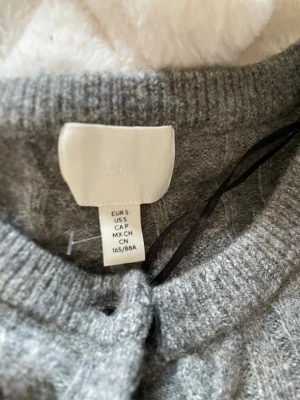 Grå kabelstickad kofta från H&M - Grå kabelstickad kofta från H&M med rund halsringning och knäppning framtill. Koftan har långa ärmar och ribbade muddar vid ärmslut och nederkant. Perfekt för lager-på-lager och enkel att matcha med olika outfits. Ny utan prislapp, endast prövad