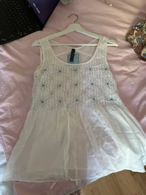 Vit somrig topp med broderi - Luftig vit topp från BL med blått broderat blommönster på överdelen. Klänningen är ärmlös och har en lätt, flowy passform med knytband i ryggen. Säljer då den är för stor för mig! Är man skicklig med att sy kan man korta ner den lite ifall man gillar den stilen mer!
