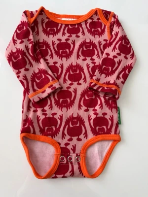 Rosa body med monstertryck Vossatassar - Säljer en rosa långärmad body från Vossatassar med coolt rött monstertryck och orange kantband. Bodyn har tryckknappar i grenen för enkel påklädning. Perfekt för dig som gillar färgglada och lekfulla plagg till de minsta.