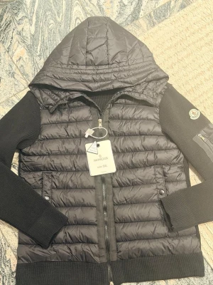 Svart pufferjacka från Moncler - Svart pufferjacka från Moncler med stickade ärmar och huva. Jackan har dragkedja framtill, två fickor med knappar och en extra ficka med dragkedja på ärmen. Moncler-logga på vänster ärm. Perfekt för kalla dagar. DET STÅR 5XL MEN DET ÄR M ❗️❗️❗️❗️