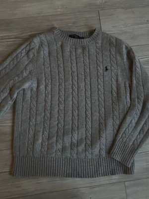 Grå kabelstickad tröja Polo Ralph Lauren - Kabelstickad grå tröja från Polo Ralph Lauren med rund halsringning och broderad logga på bröstet. Tröjan har långa ärmar och breda ribbade muddar vid ärmslut och nederkant. Perfekt för lager-på-lager och klassisk stil.
