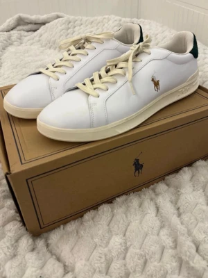 Vita Polo Ralph Lauren sneakers - Snygga vita sneakers från Polo Ralph Lauren med klassisk logga på sidan och mörkgrön mockadetalj vid hälen där det står 'Polo'. Skorna har en krämfärgad sula och snören. Perfekta för en stilren och sportig look. Köpta för 1200kr, aldrig använda. Pris kan diskuteras