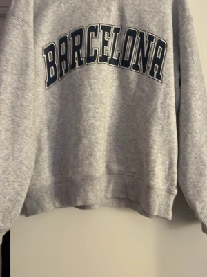 Grå sweatshirt med Barcelona-tryck - En grå sweatshirt med stort svartvitt 'BARCELONA'-tryck över bröstet. Materialet känns mjukt och bekvämt.