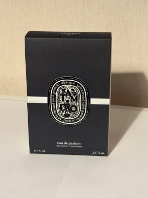 Diptyque Tam Dao Eau de Parfum 75ml - Diptyque Tam Dao Eau de Parfum i en elegant svart kartong med vit detalj och klassisk oval logotyp framtill. Parfymen kommer i 75 ml flaska och är känd för sina träiga och fräscha doftnoter. Perfekt för dig som gillar exklusiva och unika dofter.