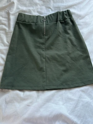 Olivgrön kjol med dragkedja framtill - Snygg olivgrön kortkjol i mjukt material med en cool silverfärgad dragkedja framtill. Kjolen har en enkel, rak passform och bred linning som ger en avslappnad vibe. Perfekt att matcha med en crop top eller hoodie. storleken funkar på de som har större storlekar än 146/152. jag har storlek XS/S och den passar mig.