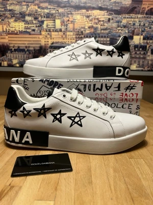 Dolce & Gabbana vita sneakers stjärnor - Snygga vita sneakers från Dolce & Gabbana med svarta stjärnor på sidorna och svart detalj med logga på hälen. Skorna har vita snören, svart hälparti och är tillverkade i slätt läder. Perfekta för dig som vill sticka ut med en cool och trendig look.