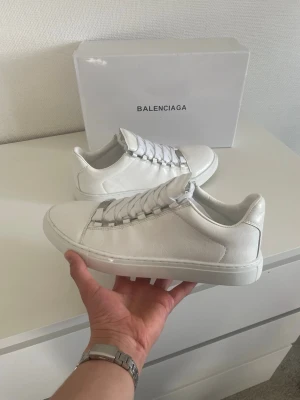 Vita Balenciaga sneakers i läder - Stilrena vita sneakers från Balenciaga i äkta läder med klassisk låg siluett. Skorna har vita snören och diskret branding, perfekt för en clean och modern look. Kommer med originalkartong.