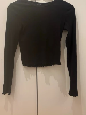 Svart croppad långärmad topp - Svart långärmad topp med croppad passform och vågig kant nertill och öppen rygg. Toppen har en öppen rygg som ger en snygg detalj. Perfekt att styla med jeans eller kjol för en trendig look.