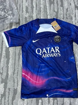 PSG tröja - Säljer en blå PSG matchtröja från Nike med Qatar Airways-logga på bröstet. Tröjan har ett galaktiskt mönster i blått, rosa och vitt, korta ärmar och klubbmärke på vänster sida. Perfekt för fotbollsfans som vill sticka ut på planen.
