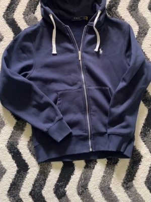 Ralph Lauren zip hoodie - Snygg mörkblå hoodie från Polo Ralph Lauren med dragkedja, vit broderad logga på bröstet och vita snören i huvan. Mjuk bomullskänsla och känguruficka framtill. Perfekt för en chill och stilren look.