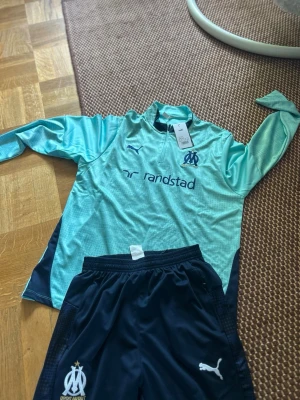 Marseille träningsset från Puma - Säljer ett snyggt träningsset från Olympique de Marseille och Puma. Tröjan är ljusblå med mörkblå detaljer, lång ärm och halv dragkedja. Byxorna är mörkblå med klubbmärke och Puma-logga, samt resår i midjan. Perfekt för fotbollsträning eller chill.