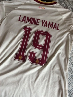 Spanien Lamine Yamal matchtröja - Officiell Spanien fotbollströja med Lamine Yamal och nummer 19 på ryggen. Tröjan är vit med vinröda detaljer och tryck, samt gulddetaljer i kragen. Tillverkad i lätt och ventilerande material, perfekt för match eller träning.