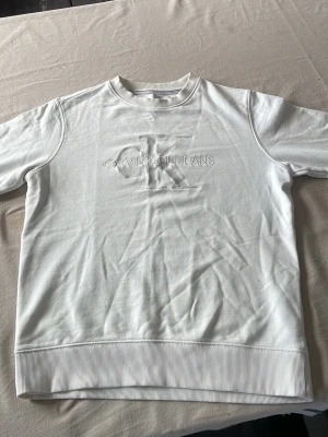 Vit sweatshirt från Calvin Klein Jeans - Vit sweatshirt från Calvin Klein Jeans med broderad logotyp på bröstet. Tröjan har rund halsringning, ribbade muddar och är tillverkad i mjukt bomullsmaterial. Perfekt för en stilren look.