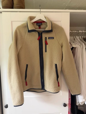 Beige fleecejacka från Patagonia - Säljer en beige fleecejacka från Patagonia med marinblå detaljer och röda dragkedjor. Jackan har hög krage, hel dragkedja framtill och fickor med dragkedja. Perfekt för lager-på-lager och chill vardagsstil. Ny pris 1 595kr.