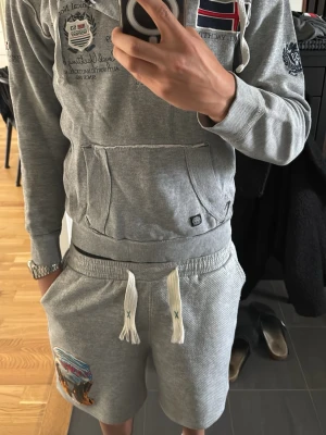 Grå hoodie från Geographical Norway - Snygg grå hoodie från Geographical Norway med broderade detaljer, norska flaggan och text på bröstet. Hoodien har huva med dragsko, knappar vid halsen och en stor magficka. Perfekt för en avslappnad och sportig stil.