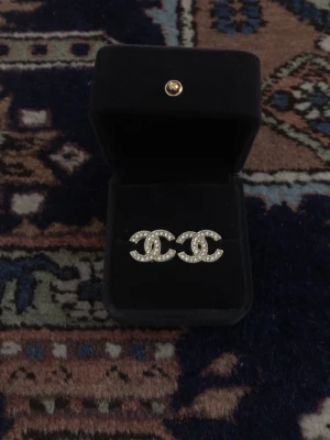 Guldiga C Örhängen med strass - Snygga C örhängen i klassisk CC-logga, täckta med glittrande strass. Örhängena är guldiga och kommer i en svart ask. Perfekta för dig som vill ha en lyxig och trendig accessoar.