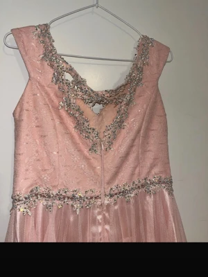 Rosa långklänning med spets och pärlor - Elegant rosa långklänning med v-ringad hals och vacker spetsdetalj. Klänningen har pärlor och broderade blommor runt halsringningen och midjan. Materialet är troligtvis polyester eller liknande, och kjolen har ett lätt, svepande fall.