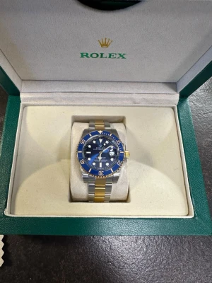 Role Submariner blå/guld herrklocka - Exklusiv Role Submariner herrklocka med blå urtavla och vridbar blå bezel. Armband i kombinerat guld och silverfärgat stål. Klassisk design med tydliga index och datumfönster. Perfekt för dig som vill sticka ut med en ikonisk accessoar.