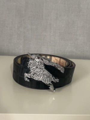 Inverterbart burberry bälte - Snyggt svart bälte med rutigt mönster och en stor, detaljerad silverspänne i form av en riddare på häst. Perfekt accessoar för att lyfta din outfit med en unik touch. Bältet har klassisk design och passar till många olika stilar.
