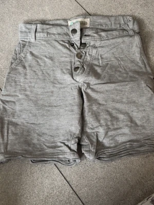 Grå shorts från AKN med knappar - Grå shorts från AKN med flera knappar framtill och bekväm passform. Gjorda i mjukt bomullsmaterial, perfekta för varma dagar. Klassisk design utan mönster och med normal midja.