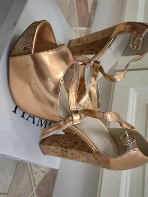 Metallic roséguldiga platåpumps 38 - Snygga platåpumps i metallic roséguld med bred klack i kork och öppen tå. Skorna har remmar över foten och spänne vid ankeln. Perfekta för dig som vill sticka ut med en trendig och glamorös look.