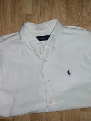 Vit skjorta från Ralph Lauren - Klassisk vit skjorta från Ralph Lauren med button-down krage och blå broderad logga på bröstet. Skjortan har långa ärmar, knäppning framtill och är tillverkad i bomull. Perfekt för en stilren och tidlös look.