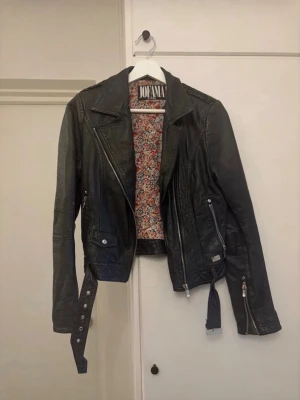 Svart skinnjacka från Jofama By Kenza - Snygg svart skinnjacka från Jofama by Kenza med coola detaljer som snedställd dragkedja, bälte i midjan och flera fickor med dragkedjor. Insidan har ett blommigt foder som ger extra stilpoäng. 