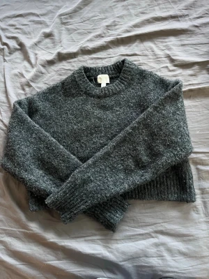 Mörkgrå stickad tröja från H&M - Mysig mörkgrå stickad tröja från H&M med rund halsringning och ribbade muddar vid ärmslut och nederkant. Tröjan har en fluffig och mjuk känsla, perfekt för kyliga dagar. Enkel och stilren design som passar till det mesta.
