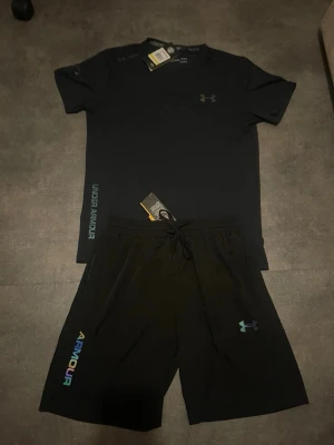 Svart träningsset från Under Armour - Snyggt svart träningsset från Under Armour med t-shirt och shorts. T-shirten har logga på bröstet och text längs sidan, tillverkad i funktionsmaterial. Shortsen har dragsko i midjan och färgglad logga på benet. Perfekt för gym eller löpning.