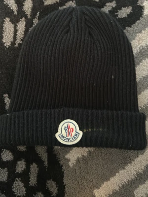 Svart Moncler mössa i ribbstickad ull - Svart ribbstickad mössa från Moncler med uppvikt kant och klassisk Moncler-logga framtill. Snygg och enkel design som passar till det mesta. Tillverkad i mjuk ull för extra värme under kalla dagar.