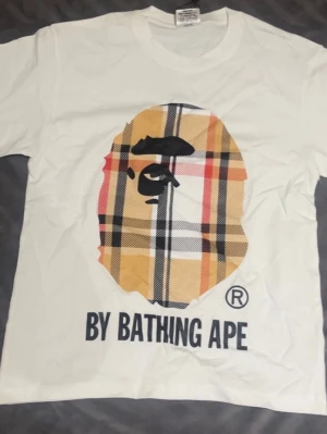 Vit BAPE t-shirt med rutig logga - Vit t-shirt från A Bathing Ape med stor rutig BAPE-logga i gult, svart, rött och vitt på bröstet. Klassisk rund hals och korta ärmar. Tryckt text 'BY BATHING APE' nedanför loggan. Perfekt för dig som gillar streetwear och unika prints.
