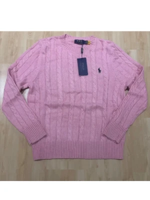 Rosa kabelstickad tröja Polo Ralph Lauren - Kabelstickad rosa tröja från Polo Ralph Lauren med rund halsringning och broderad logga på bröstet. Tröjan har ribbade muddar och är perfekt för en snygg och klassisk look.