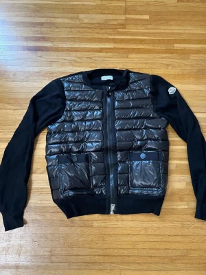 Svart cardiganjacka från Moncler - Snygg svart cardiganjacka från Moncler med quiltad, glansig front och två fickor med knappstängning. Ärmar och rygg i mjukt stickat material, dragkedja framtill och klassisk Moncler-logga på ärmen. Perfekt för lager på lager och streetstyle.