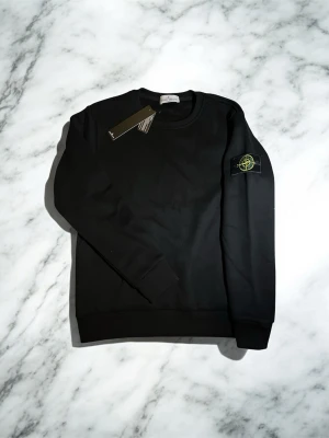 Svart sweatshirt från Stone Island - Säljer en svart sweatshirt från Stone Island med klassisk rund hals och ribbade muddar. Tröjan har den ikoniska Stone Island-loggan på vänster ärm och är tillverkad i mjukt bomullsmaterial. Perfekt för en clean och stilren look.