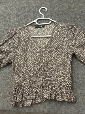 Blommig v-ringad blus från Bik Bok - Söt blommig blus från Bik Bok med v-ringning och korta puffärmar. Blusen har volangkant nedtill och smockad midja för en feminin siluett. Perfekt till jeans eller kjol för en trendig look.