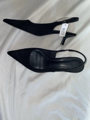 Svarta spetsiga pumps från H&M - Snygga svarta pumps från H&M med spetsig tå och smal klack. Skorna har en öppen häl med justerbar rem och är tillverkade i mockaimitation. Perfekta för dig som gillar en stilren och trendig look.