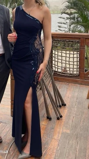 Marinblå oneshoulder långklänning - Elegant marinblå långklänning med oneshoulder-design och transparent padded spetsdetalj längs sidan. Klänningen har en hög slits framtill och glittriga paljetter längs kanten. Perfekt för speciella tillfällen där du vill sticka ut. Använd 1 gång.