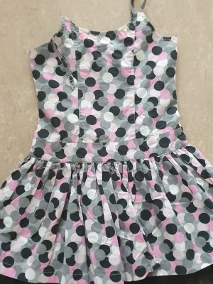 Klänning - Söt kort klänning med smala axelband och volangkjol. Prickigt mönster i svart, grått, vitt och ljusrosa. Klänningen har en smockad rygg för extra komfort och en lätt, luftig känsla. Perfekt för varma dagar och somriga vibes. Ena axelbandet har gått av.