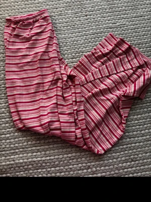 Randiga mjukisbyxor från Gina Tricot - Säljer ett par mysiga mjukisbyxor från Gina Tricot i storlek XS. Byxorna har rosa, röda och vita ränder, resår i midjan och dragsko för bästa passform. Perfekta för chill hemma eller som pyjamasbyxor. Materialet är mjukt och skönt mot huden.