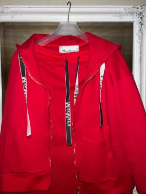 Röd hoodie och kjol  från Max Mara med dragkedja - Snygg röd hoodie och kjol set från Max Mara med dragkedja framtill och vita snören med svart text. Hoodien har fickor på sidorna och en klassisk huva. Tillverkad i mjukt material som känns skönt mot huden. Perfekt för en avslappnad och trendig look. Storlek är XL men passar bra även på M L
